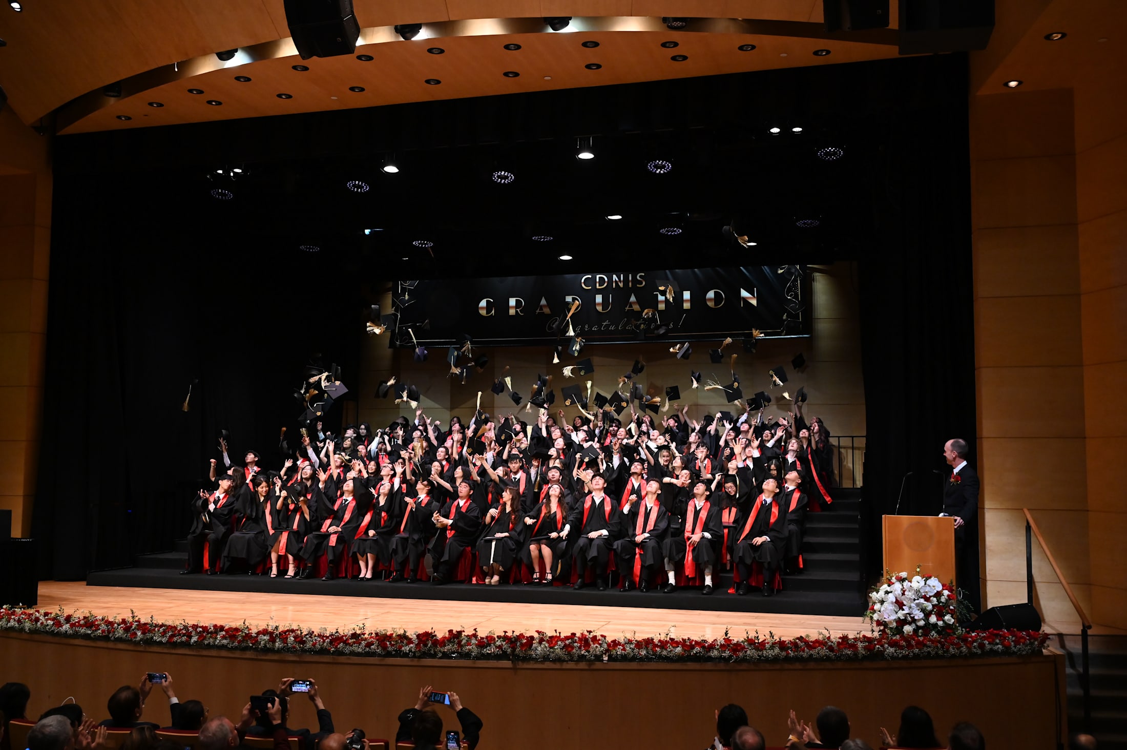 A large group of people in red robes and gowns are gathered on a stage, with a backdrop displaying the words %22Commencement%22 and a podium in the foreground.